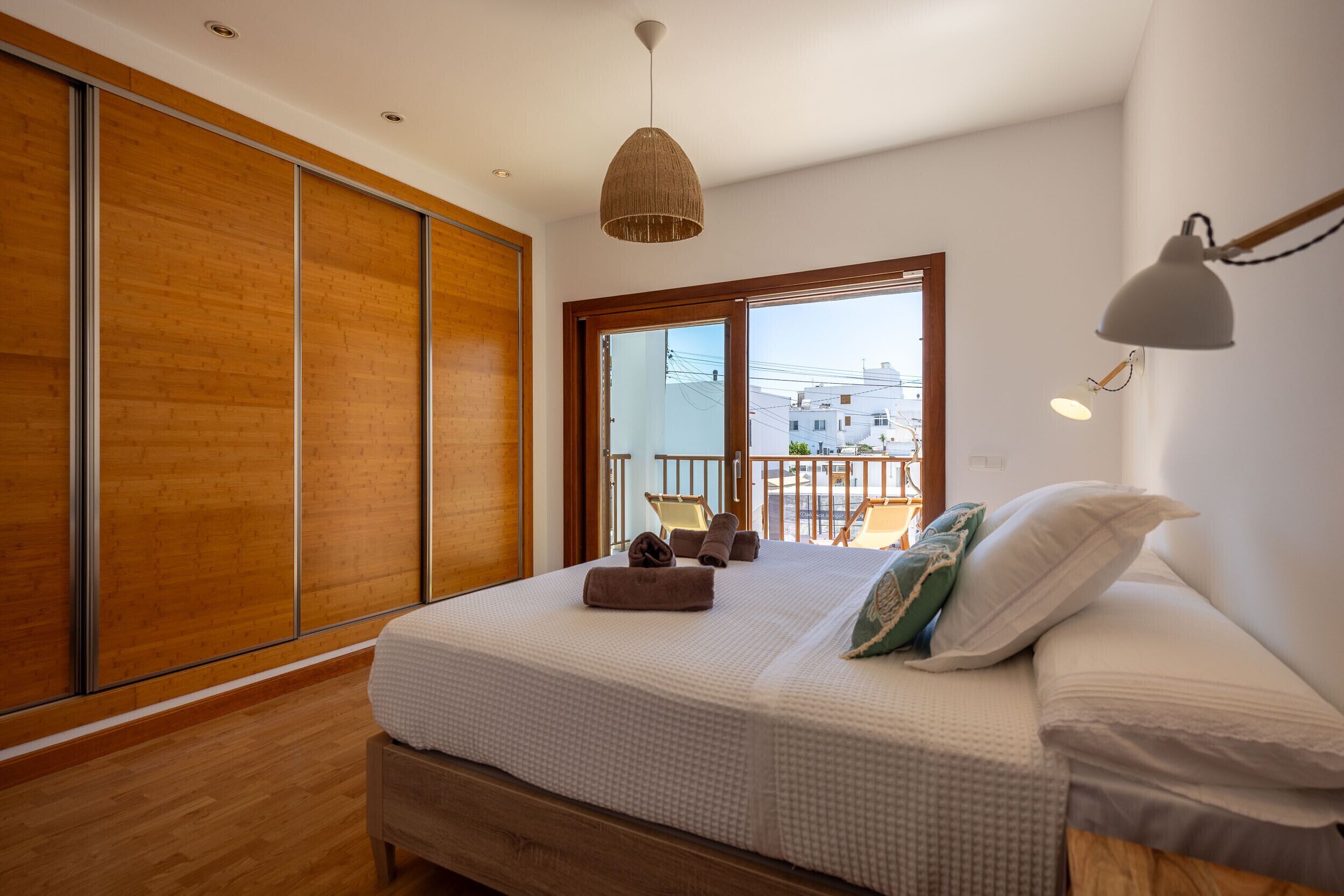 A bedroom with a large bed, wooden flooring, a balcony with chairs, and a view of buildings.