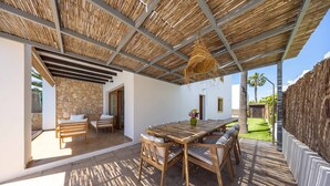 Eine Terrasse im Freien mit Holztisch und -stühlen, einer Steinmauer und einem strohgedeckten Dachaufbau.