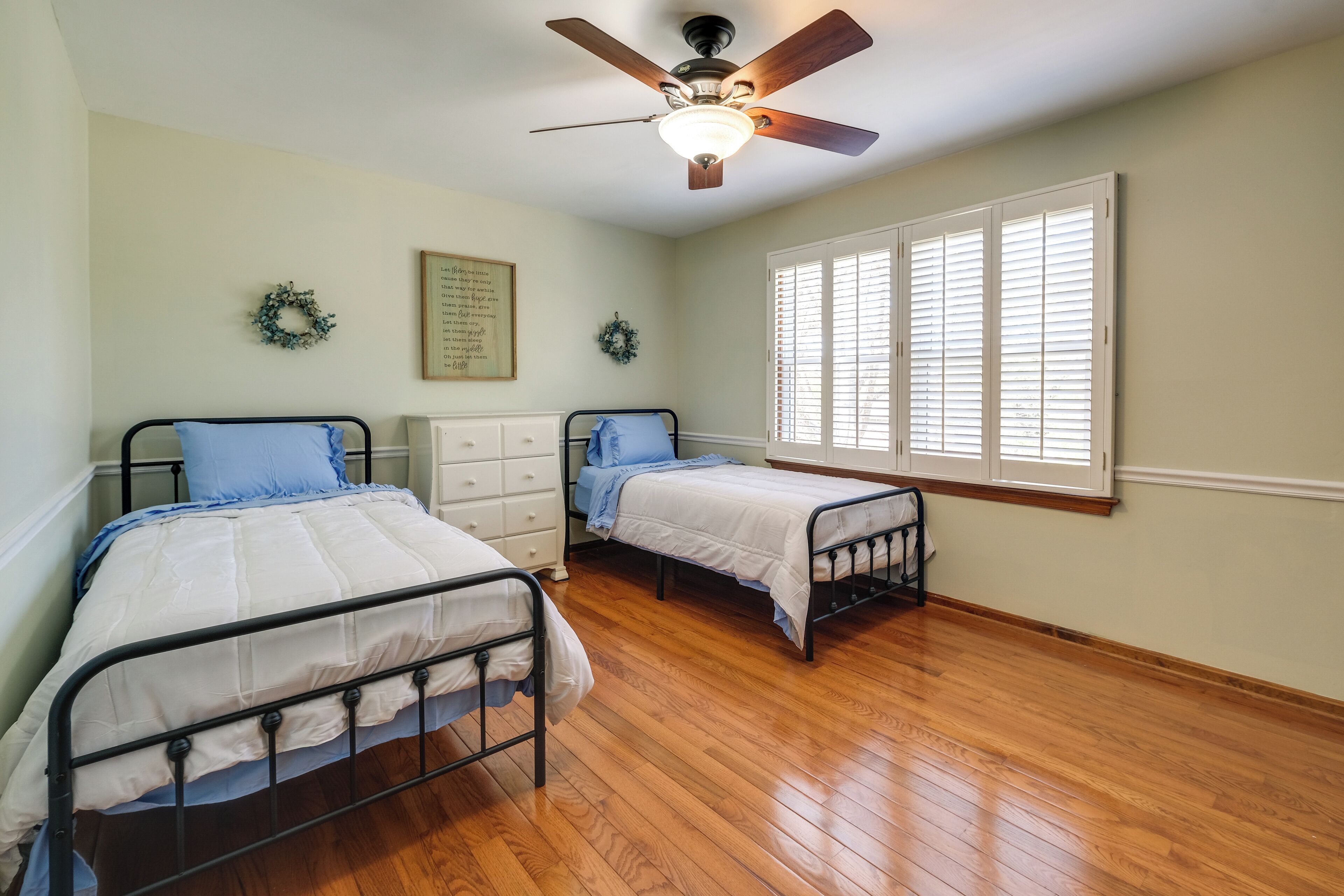 Quarto com duas camas, um ventilador de teto, uma janela com persianas e uma cômoda.