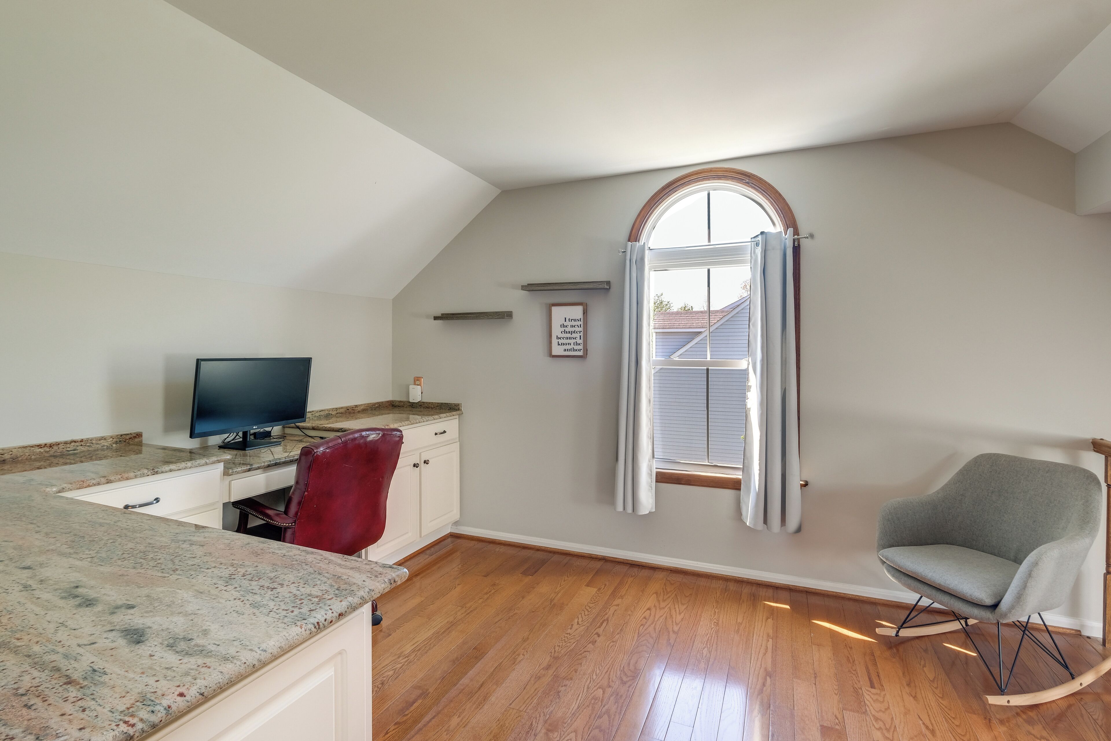 A room with a wooden floor, a desk with a computer, a rocking chair, and a window with curtains.