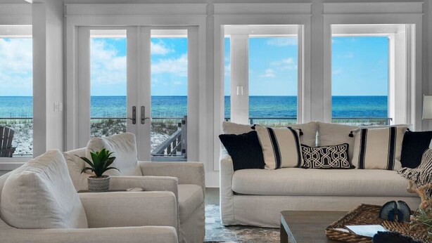 Ein geräumiges Wohnzimmer mit einem großen Fenster mit Meerblick, ein beiges Sofa mit gemusterten Kissen und eine kleine Pflanze auf einem Beistelltisch.