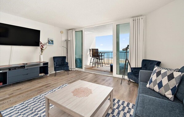 A modern living room with a flat-screen TV, a sofa, a coffee table, and a balcony with a view of the sea. - Beachfront 1 BR 1.5 BA Unit with a Pool and Fantastic Ocean Views! (Ocean City)