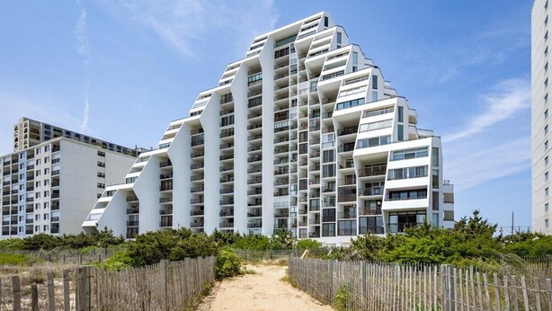 Un bâtiment moderne à plusieurs étages, doté d’un design anguleux original, situé près d’un sentier sablonneux bordé d’une clôture en bois.