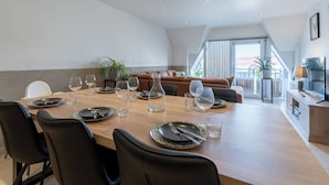 Uma área de refeições com uma mesa comprida posta para seis pessoas, rodeada por cadeiras pretas, um sofá de couro marrom e uma varanda ao fundo.
