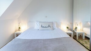 Quarto com teto inclinado, cama com travesseiro que diz 'praia', mesas de cabeceira com abajures e um espelho.