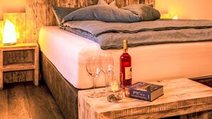 Un lit avec une tête de lit en bois, des oreillers, une bouteille de vin, des verres et un livre sur une table de chevet en bois.
