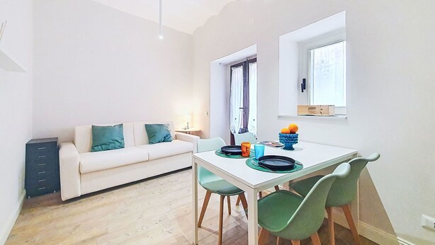 A modern living room with a white sofa, a dining table with chairs, and a window with a view.