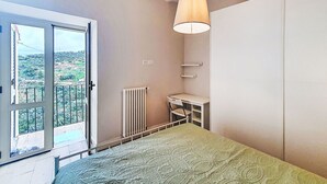 A bedroom with a bed, a desk, a chair, a radiator, a window, and a view of the outdoors.