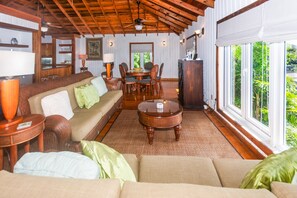A spacious living area with a wooden ceiling, a dining table with chairs, and large windows offering a view of greenery.