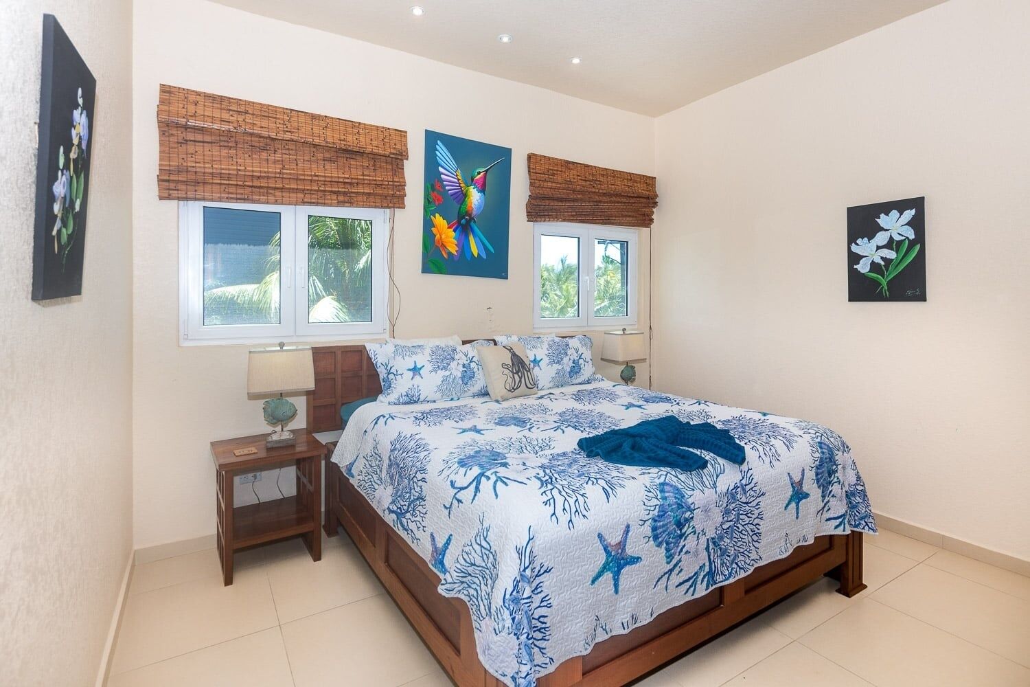 A bedroom with a bed, two windows with blinds, a nightstand, a lamp, and wall art.