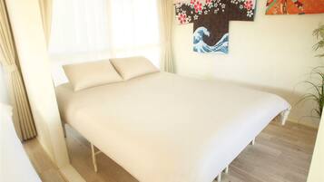 A bed with a white mattress and pillows, a wooden floor, a window with sheer curtains, and a wall with a framed artwork.