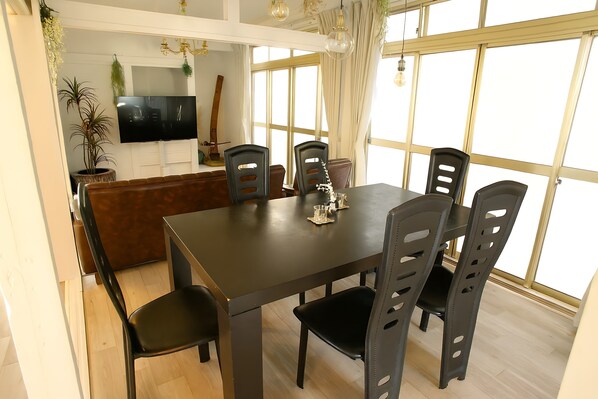 A dining area with a dark wooden table and six chairs, a TV mounted on the wall, and a large window with curtains.