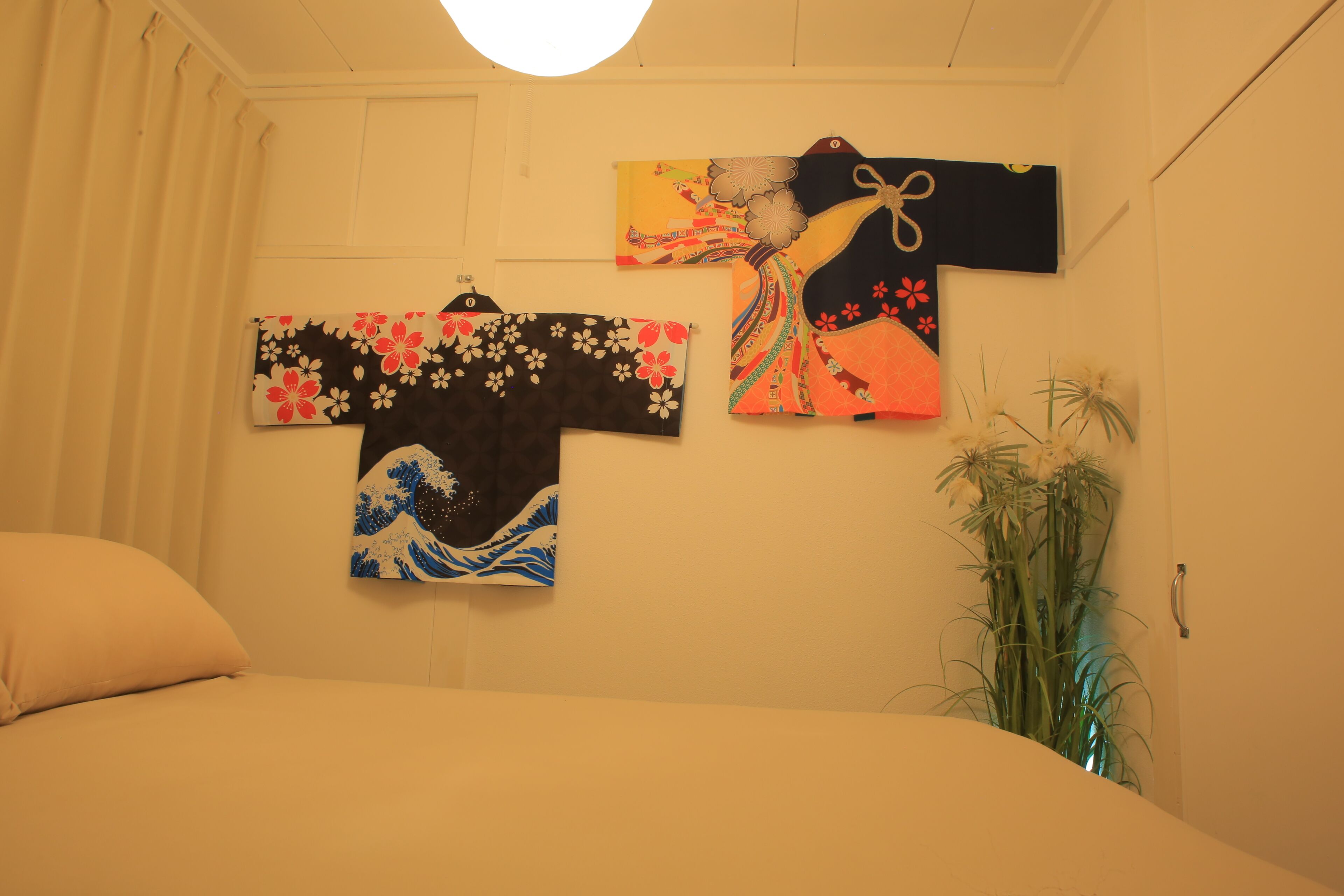 A room with a bed, a wall-mounted Japanese garment, and a potted plant.