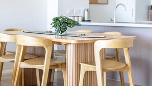 A modern kitchen with a central island, wooden chairs, a potted plant, and a wall clock. - Beachside Bliss 3BDR mins to Caves Beach & Bistros (Caves Beach)