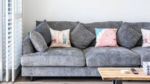 A grey sofa with multiple cushions, a wooden coffee table, and a surfboard mounted on the wall. - Beachside Bliss 3BDR mins to Caves Beach & Bistros (Caves Beach)