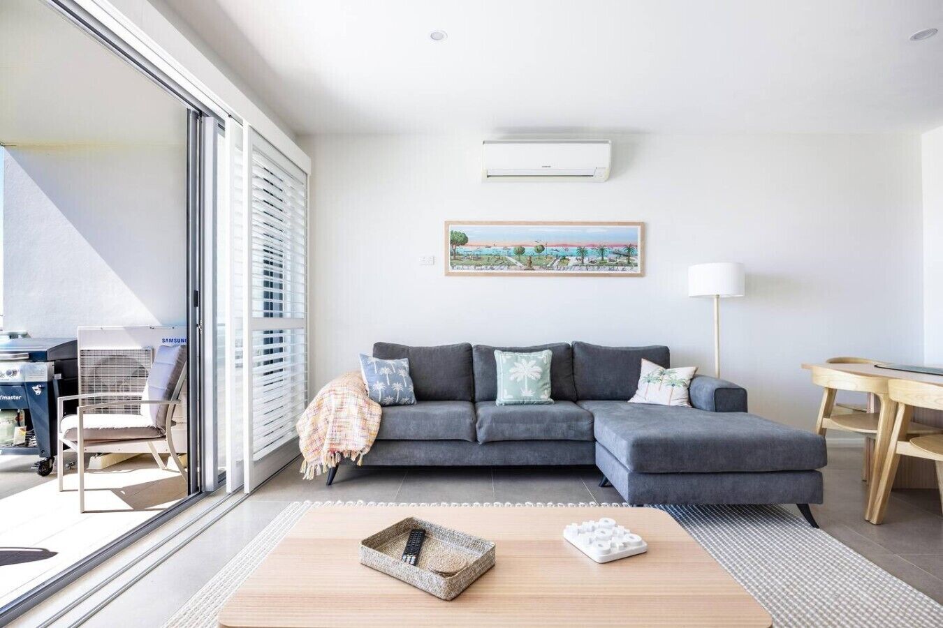 A modern living room with a grey sectional sofa, a large window with blinds, a painting on the wall, and a small table with a lamp.