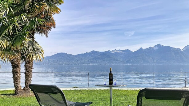 Zwei Stühle, ein Tisch mit einer Flasche und Gläsern sowie der Blick auf einen See mit Bergen im Hintergrund.