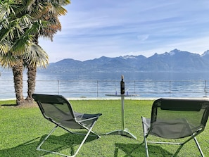 Due sedie, un tavolo con una bottiglia e dei bicchieri, e vista su un lago con montagne sullo sfondo.