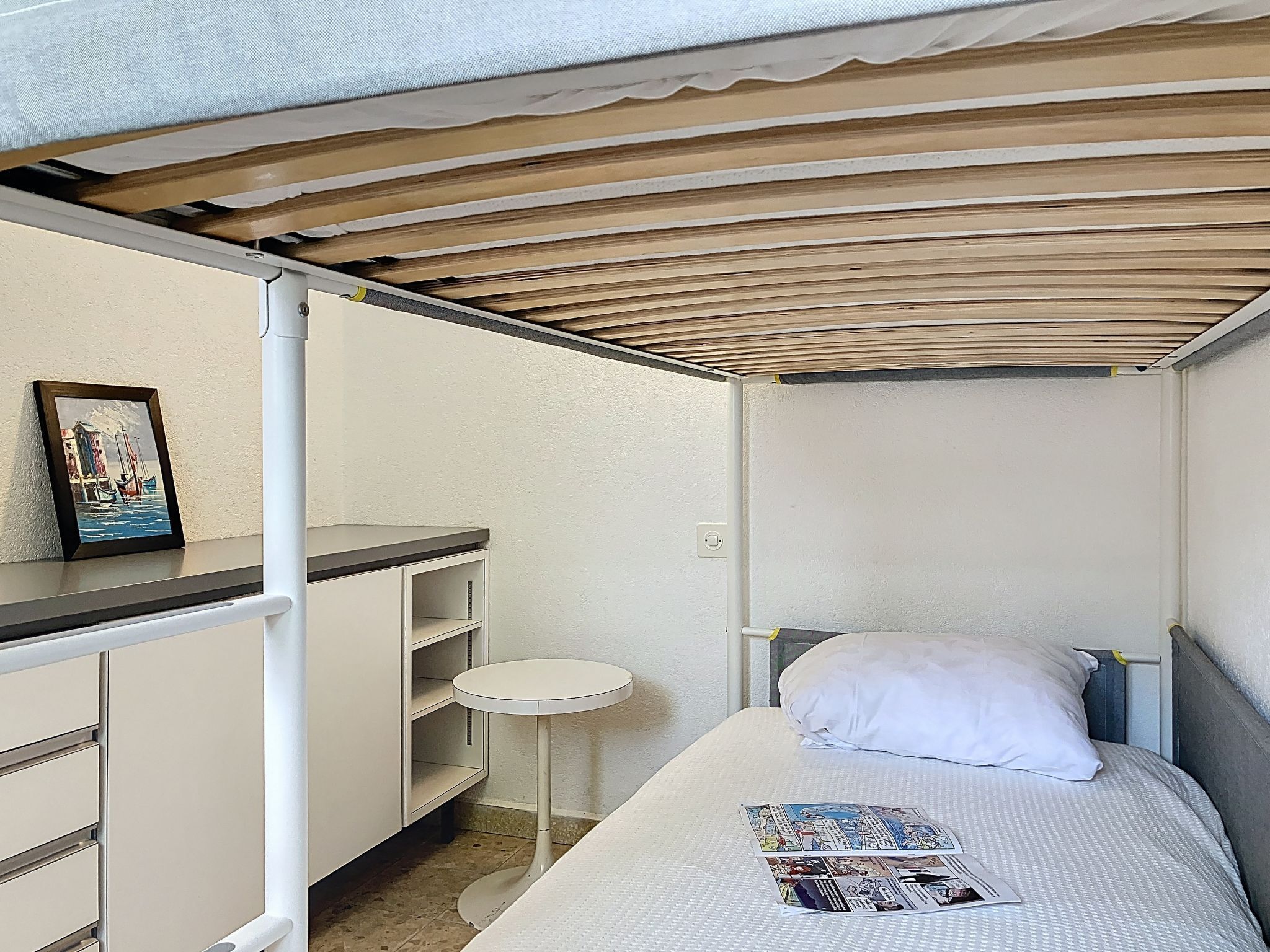 A compact room with a bed, a small table, a shelf unit, and a framed picture on the wall.