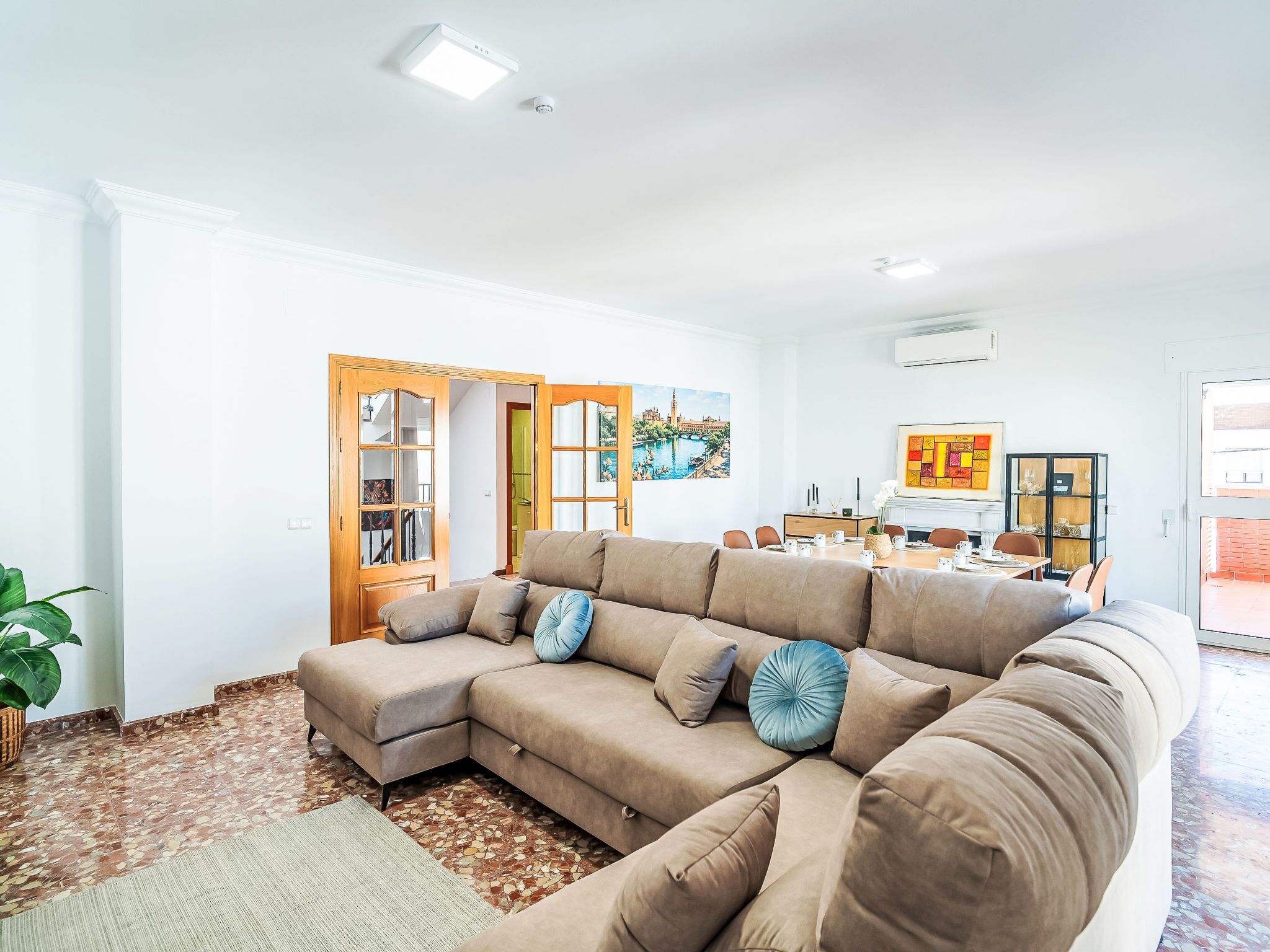 A living room with a sectional sofa, a dining area with a table and chairs, a large window with a view, and a door leading to another room.