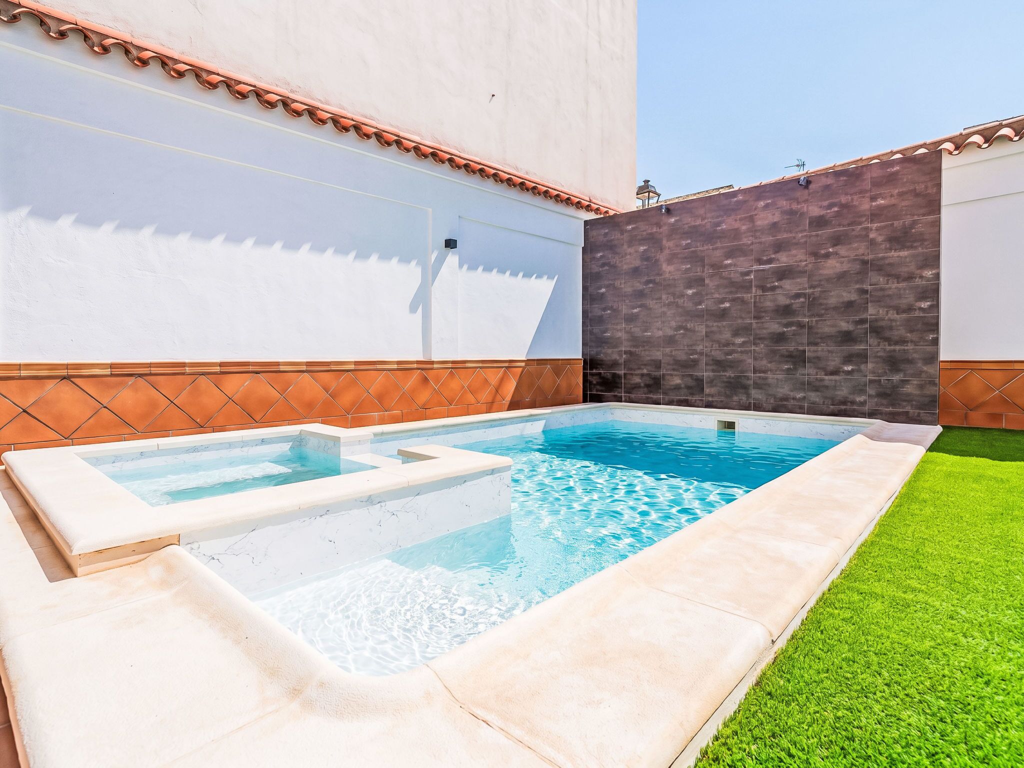 Una piscina con un jacuzzi pequeño, rodeada por un muro blanco y un muro con azulejos oscuros.