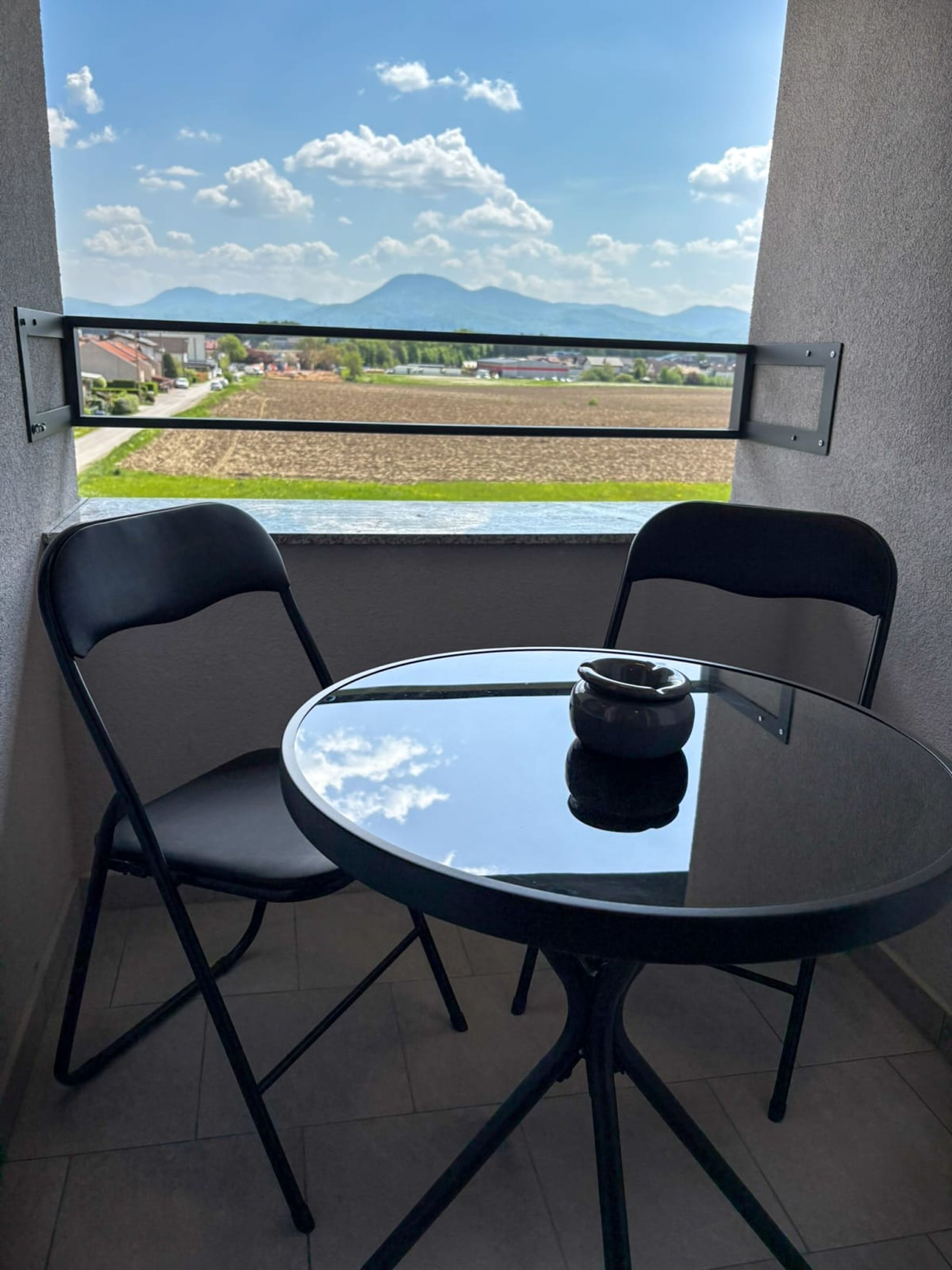 Un balcón con dos sillas y una mesa, vista a un campo y montañas a lo lejos.