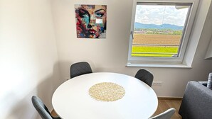 A dining area with a round table, four chairs, an air conditioner, a window with a view of fields, and a colorful abstract painting on the wall. - Southwest Ljubljana Private Parking 8 (Ljubljana)