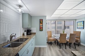 Una cocina con encimeras de granito, lavamanos de acero inoxidable y una cafetera. Hay un comedor con sillas de mimbre y una mesa.