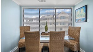 A dining area with wicker chairs and a glass table, large windows with blinds, and a painting on the wall. - New to VRBO 3 Bedroom Villa - Golf cart included - 604 Newhaven (Fripp Island)