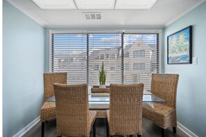 Un comedor con sillas de mimbre y una mesa de vidrio, ventanas amplias con persianas, y un cuadro en la pared.