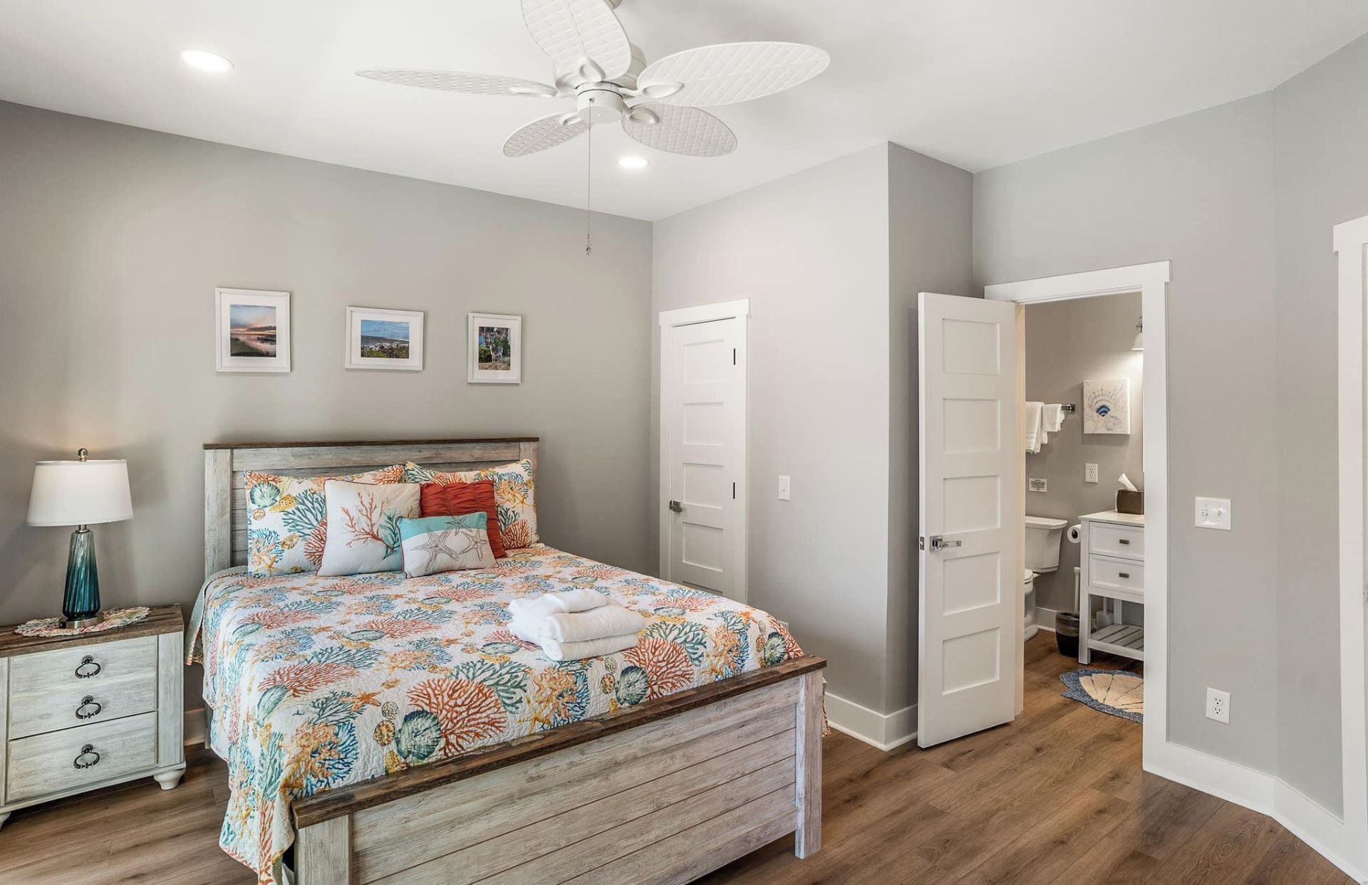Une chambre à coucher avec un lit, une table de chevet, une lampe, un ventilateur de plafond et une salle de bain visible à travers une porte ouverte.