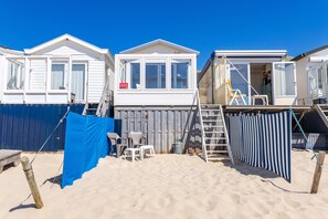 Ein Ferienhaus am Strand mit einer Veranda, einer kleinen Terrasse und einer sichtbaren Treppe zum Obergeschoss.
