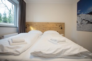 A double bed with white linens and towels, a wooden headboard, a nightstand with a lamp, and a window with curtains.