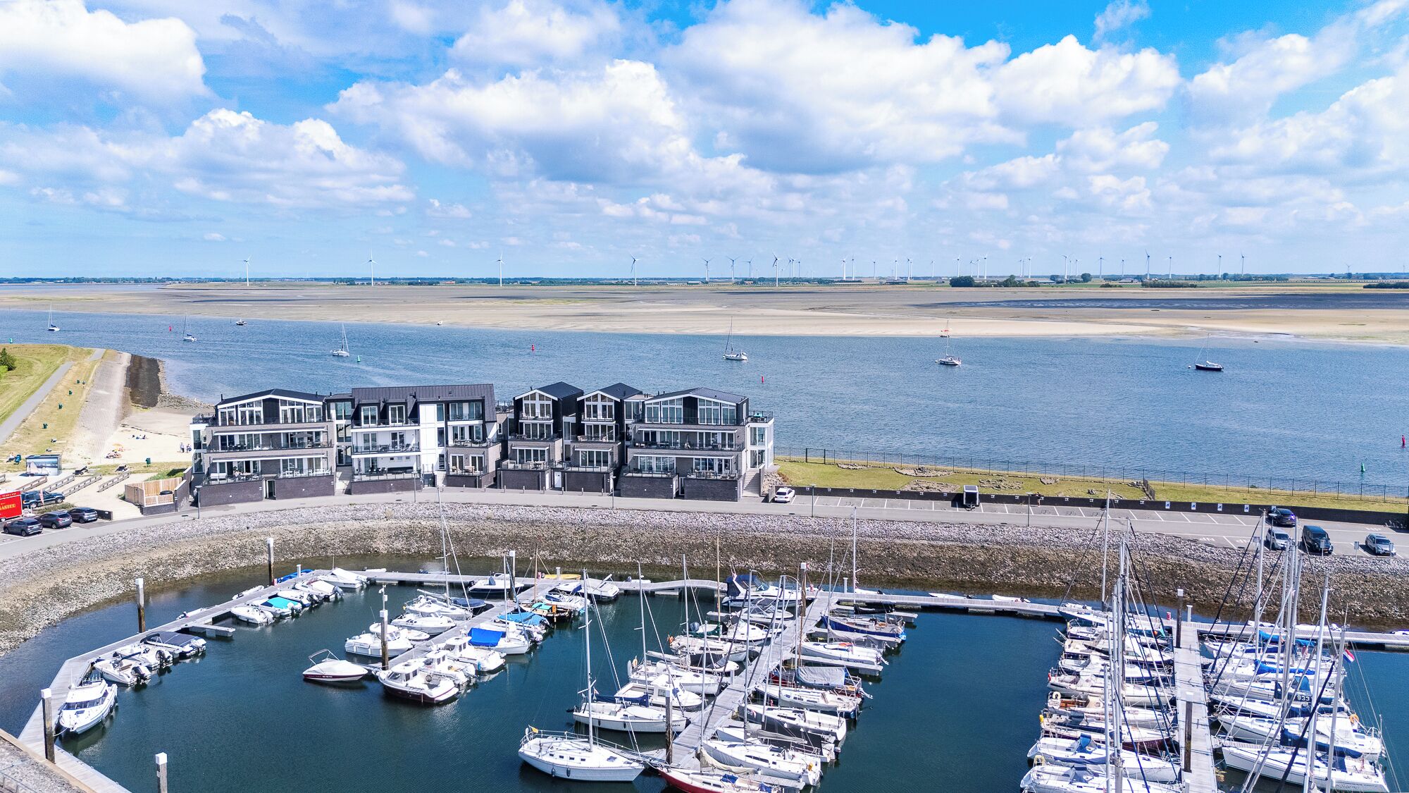 Een jachthaven met aangemeerde boten, een gebouw met meerdere verdiepingen en een strand op de achtergrond.