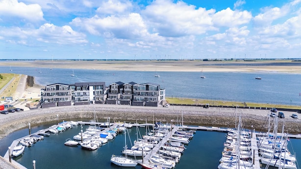 A marina with boats docked, a building with multiple levels, and a beach in the background.
