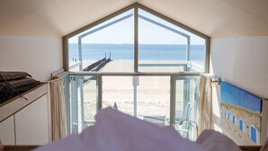 A room with a large window overlooking a beach, a bed, and a painting on the wall. - Slaapstrandhuisje - Strand Dishoek 37 I Dishoek (Koudekerke)