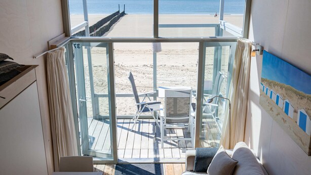 A room with a large glass door leading to a beach view, a chair, a small table, and a painting on the wall. - Slaapstrandhuisje - Strand Dishoek 37 I Dishoek (Koudekerke)
