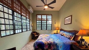 Un dormitorio con cama, una silla, un ventanal, un ventilador de techo y una lámpara.