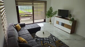 Een moderne woonkamer met een hoekbank, een salontafel, een televisie op een houten kast en een groot raam met jaloezieën.