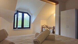 Een slaapkamer met schuine wand, een raam met uitzicht op bomen, een bed met witte lakens en een kledingkast.