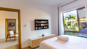 Hotellihuone, jossa on sänky, seinälle asennettu televisio, puinen penkki ja näkymä palmuille ikkunasta.
