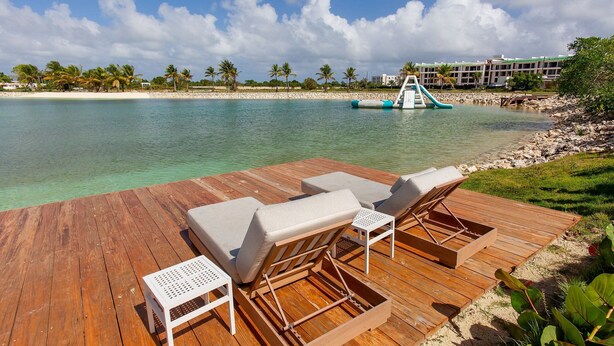 A wooden deck with lounge chairs and a small table, overlooking a clear turquoise sea with a palm tree-lined beach and a water slide in the background. - 1BR, artificial beach, pools, gym, location (Punta Cana)