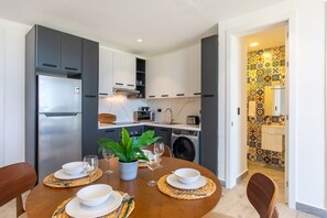 A dining area with a table set for four, a refrigerator, a washing machine, and a kitchen with white cabinets and a patterned backsplash. - 1BR, artificial beach, pools, gym, location (Punta Cana)