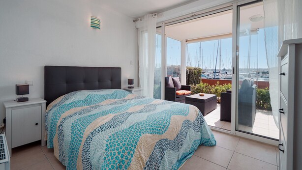 A bedroom with a large bed, a nightstand, a chair, and a view of a marina through a sliding glass door. - Quiet Elegance Near the City Center Agde (Agde)