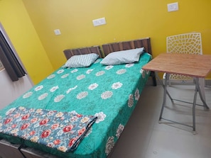 Una habitación con cama, una mesita y una silla. Hay dos enchufes en la pared.