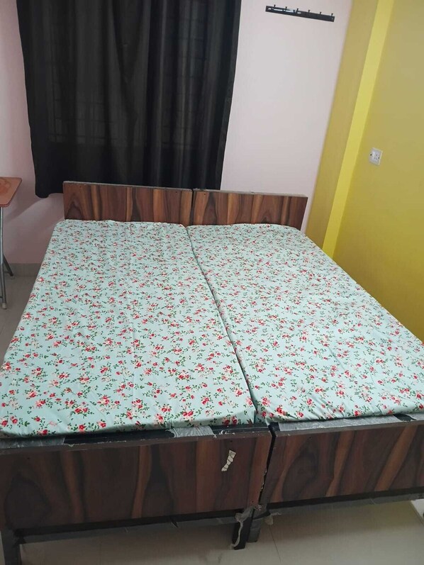 Una cama con colcha estampada, cabecera de madera, cortina negra y pared amarilla.