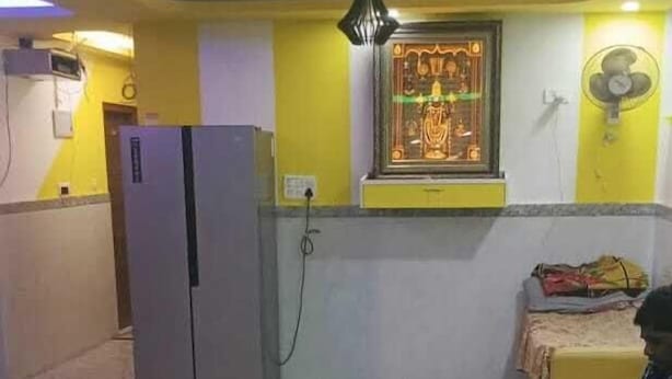 A room with a refrigerator, a fan, a small shrine, and a person sitting on a bench. - Rudra Co-Living Hotel-PG (Bengaluru)