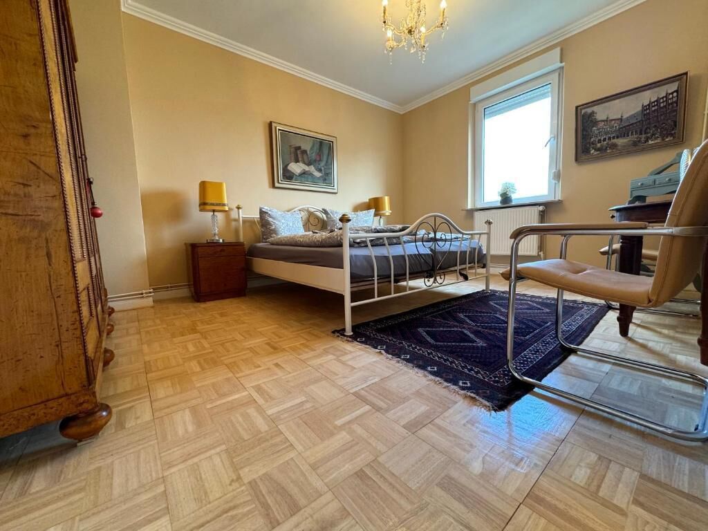 A bedroom with a bed, bedside tables, a chair, a chandelier, a window, and a painting on the wall.