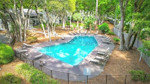 A swimming pool surrounded by lounge chairs and trees, with a fence and a building in the background. - Stones Throw to the Shore (Hilton Head Island)