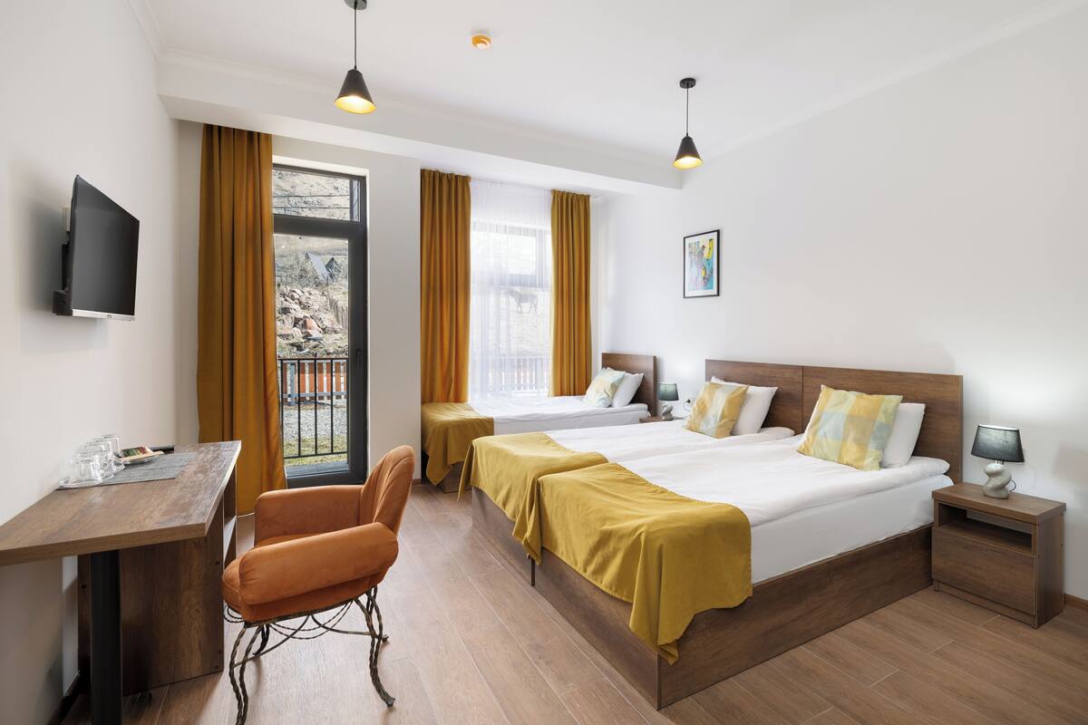 A hotel room with a bed, two bedside tables, a desk with a chair, a TV, and a window with curtains.